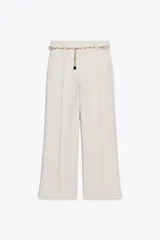 Pantalón tobillero de tiro alto color crudo, con pernera recta y cinturón de cadena dorada con dije circular negro.