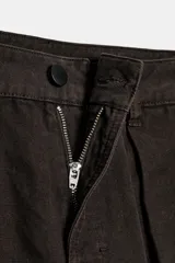 Pantalón cargo de algodón color marrón oscuro, tiro medio, con pliegues frontales, bolsillos delanteros ocultos y bolsillos de plastrón con solapa y botones a presión en las piernas. El bajo del pantalón tiene una trabilla con botón y se muestra doblado hacia afuera.