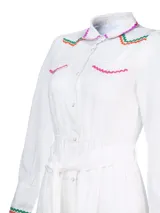 Vestido camisero largo de lino blanco con cuello camisero, cierre frontal con botones y cinturón de lazo. Presenta detalles de bordado ondulado en verde, naranja y rosa en cuello, bolsillos, puños y ruedo.