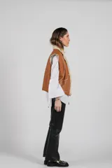 Chaleco reversible de piel de cordero color beige claro, con forro de cuero marrón visible en el borde. Se lleva sobre una camisa blanca de manga larga.