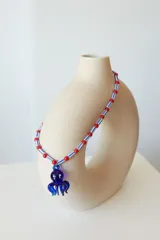 Collar de cuentas de plástico verdes y blancas alternadas, con dije de pulpo rojo de vidrio. El dije de pulpo tiene ojos blancos con pupilas negras.