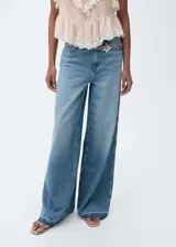 Pantalón de jean celeste de corte wide leg, tiro alto, con cinco bolsillos, cierre con cremallera y botón, trabillas y bajo deshilachado. Incluye un cinturón de cadena plateada con dije de flor.