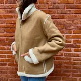 Campera de cuero ovino color tostado con interior de piel sintética blanca. Tiene cuello alto con solapas y cierre frontal con cremallera.