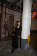 Vestido largo negro de tela elastizada con escote profundo, frunces y falda con vuelo. Tiene mangas largas y cierre invisible en la espalda.