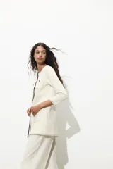 Chaqueta color crema de punto con cuello redondo y manga larga. Tiene bolsillos delanteros de plastrón, pespuntes combinados a contraste y cierre frontal con botones dorados.