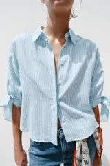 Camisa celeste y blanca a rayas verticales, confeccionada con lino e hilo metalizado. Tiene cuello solapa, mangas 3/4 con lazos en los puños y cierre frontal con botones ocultos por solapa.