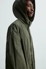 Parka verde militar de tejido técnico ligero, corte oversize, cuello alto con capucha ajustable, manga larga con puños elásticos, cintura ajustable con cordón interno, bolsillos con solapa, bajo asimétrico con abertura trasera ajustable y cierre frontal con cremallera oculta y botones a presión.