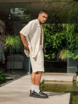 Camisa de manga corta color crudo/blanco, confeccionada en tejido de punto abierto con textura calada y cuello polo. Presenta cierre frontal de botones.