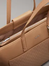 Cartera tote Calvin Klein color tostado, de tamaño mediano, confeccionada en cuero sintético granulado con textura de logo monograma en el bolsillo delantero. Tiene asas fijas, cierre magnético, bolsillo exterior, compartimento interior con cremallera y colgante extraíble con logo.