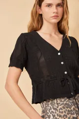 Top de tejido de punto calado color negro, con escote en V, mangas cortas con puños y cierre frontal con botones. Presenta un lazo ajustable en la cintura.