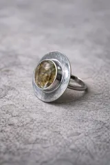Anillo artesanal de alpaca con acabado envejecido. Presenta una piedra central facetada de color ámbar/marrón claro, engastada sobre una base circular amplia y texturizada. La banda del anillo es ajustable.