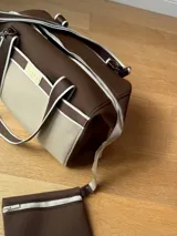 Bolso tipo tote de neopreno color negro y marrón, con cierre superior, bolsillo frontal marrón con logo rojo, bolsillo interno y externo con cierre, doble asa de mano marrón con detalles blancos y opción de correa larga.