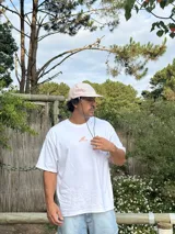 Gorro de visera curva con diseño a rayas verticales en tonos rosado pálido y blanco. La corona tiene un bordado en azul claro con texto. La visera es de color beige claro. Incluye un cordón negro ajustable para colgar del cuello.