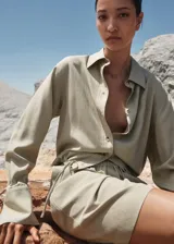 Camisa color beige de corte crop, confeccionada en mezcla de lino y lyocell. Tiene cuello camisero, cierre frontal con botones, manga larga con puños abotonados y lazo ajustable en la cintura.