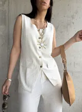 Chaleco color crema de lino con escote cuadrado, sin mangas, con cierre frontal de botones, dos bolsillos de parche y corte irregular en el ruedo.