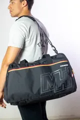 Bolso deportivo tipo duffel bag, color negro, con grandes iniciales 'MH' en el frente en tono gris oscuro. Presenta detalles de franjas horizontales en rojo, amarillo y negro sobre los cierres de los compartimentos. La correa de hombro lleva la inscripción 'NBA' en relieve.