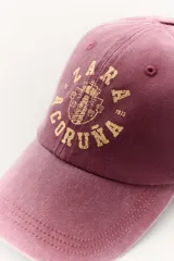 Gorra color borgoña con bordado dorado de la marca Zara, con la inscripción "Zara" y "A Coruña 1975".