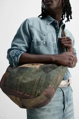 Bolso de hombro tipo bandolera con estampado de parches de denim en tonos verdes, marrones y azules. Tiene un compartimento principal con cierre de cremallera, un bolsillo interior con cremallera y un bolsillo trasero con cremallera. El asa de hombro es ajustable.