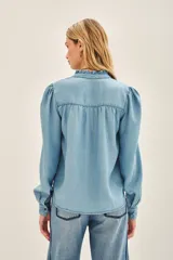 Camisa celeste de tencel con calce recto, cuello redondo con volados, canesú en delantero y espalda, y mangas largas terminadas en puño con botón snap.