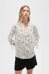Blusa color beige con estampado de arabescos blancos, cuello mao, mangas 3/4 con lazos en los puños, cierre parcial con botones y bolsillos frontales.