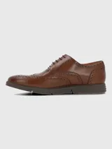 Zapato de vestir tipo Oxford o Brogue, color marrón cuero, con puntera y laterales decorados con perforaciones (broguing). Posee cordones y una suela de goma gruesa y moderna de color marrón oscuro.