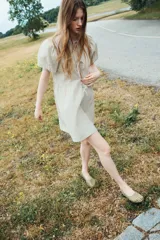 Vestido corto de algodón a cuadros en tonos beige y verde claro. Tiene cuello redondo con abertura y lazos, mangas cortas abullonadas y falda con leve vuelo.