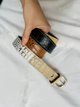 Cinto de cuero con hebilla metálica plateada y trabilla marrón. El cuero presenta diferentes texturas y colores: beige con textura de piel de reptil, negro con textura de cocodrilo, marrón con textura de piel de reptil y blanco con estampado dorado.