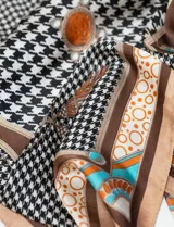 Pañuelo cuadrado de satén estampado con motivos geométricos y florales en tonos marrón, beige, naranja, celeste y negro.