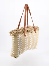 Bolso tipo tote color beige con diseño geométrico en blanco y flecos en la parte superior. Tiene doble asa de hombro marrón.