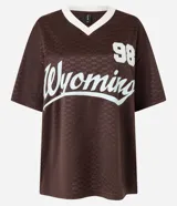 Remera marrón con diseño de damero, cuello en V con borde blanco, mangas cortas y estampado frontal con el número 98 y la palabra "Wyoming" en color celeste.
