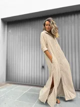 Vestido camisero largo con estampado de rayas verticales en beige y blanco. Presenta cuello camisero, cierre frontal con botones, mangas largas y tajos laterales en el ruedo.