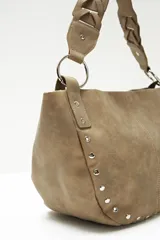 Cartera tipo hobo color beige con tachas plateadas en el borde inferior y asa de hombro trenzada.