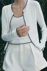 Chaqueta blanca de punto con cuello redondo y manga larga. Tiene vivos negros en el cuello, el borde frontal y los puños. Se cierra con botones en el frente.