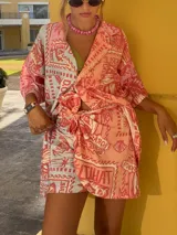 Falda pareo corta, color coral, con estampado de temática playera que incluye sellos postales, conchas marinas y texto. Se anuda en la cintura.