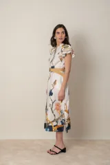 Vestido midi camisero de algodón color natural con estampado de flores, animales y ramas en tonos rojo, marrón, verde y azul. Tiene cuello mao, mangas cortas con volados, bolsillos laterales y cinturón de soga color beige.