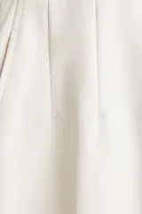 Pantalón palazzo color blanco hueso de tiro alto con elástico oculto en la cintura, cierre lateral oculto y pinzas que realzan la figura. Modelo de corte holgado con bolsillos al bies, bolsillos decorativos atrás y piernas tres cuartos con raya adelante y atrás.