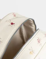 Mochila de cuerina color crema con estampado de flores bordadas, bolsillo frontal con cierre, correas regulables, manija superior, acceso con cierre superior, bolsillos laterales y tag de cuerina.