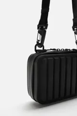 Bandolera rígida negra con acabado engomado y relieve. Tiene un compartimento principal con doble cremallera, un bolsillo interior de rejilla y asa de hombro ajustable y extraíble.