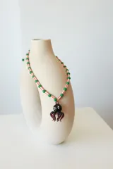 Collar de cuentas de plástico verdes y blancas alternadas, con dije de pulpo rojo de vidrio. El dije de pulpo tiene ojos blancos con pupilas negras.