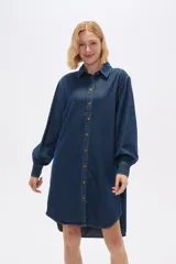 Vestido camisero de jean azul oscuro, corte holgado, largo a la rodilla, con cuello camisero, mangas largas abullonadas con puños abotonados y cierre frontal con botones.