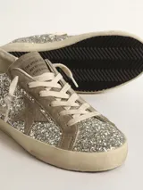 Zapatillas deportivas estilo urbano con upper completamente cubierto de glitter plateado. Presentan detalles en gamuza color arena, incluyendo la estrella lateral característica. El talón tiene un acabado metálico color cobre. La suela es de goma color beige con efecto desgastado.