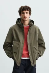 Parka verde militar de tejido técnico ligero resistente al viento, con cuello alto, capucha y manga larga con puños ajustables. Tiene bolsillos de plastrón con solapa en la cadera, bolsillo interior y bajo ajustable con elásticos. El cierre frontal es con cremallera oculta por solapa con botones a presión.