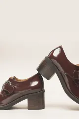 Zapato de cuero vacuno charol negro, con punta redondeada y detalle de perforado en la capellada. Tiene una tira con hebilla metálica y un taco cuadrado de 4 cm.