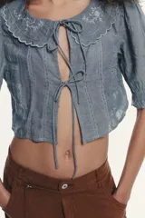 Camisa corta celeste confeccionada en algodón 100%. Presenta cuello bobo con bordados florales, mangas cortas abullonadas y cierre frontal con lazadas.