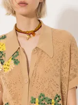 Túnica tejida color beige, con diseño calado y bordado de limones en el frente. Tiene cuello tipo camisa, mangas cortas y cierre frontal con botones.