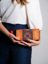 Billetera rectangular de tela bordada con motivos tribales en tonos celeste, azul, marrón y rosa. Tiene cierre con cremallera y tirador con pompón y cuentas de madera.