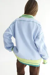 Buzo celeste con cuello polo y puños en rib color verde lima. Tiene un parche con el logo de la marca en el pecho y rayas horizontales en el ruedo.
