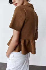 Chaqueta color marrón de tejido de punto, con cuello solapa, manga corta y bajo evasé con volantes. Cierre frontal con botones forrados a tono.