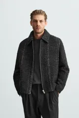 Sobrecamisa negra con textura de tejido bouclé y rayas verticales plateadas. Tiene cuello solapa, manga larga con puños abotonados, bolsillos laterales y cierre frontal con cremallera.