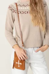 Bolso pequeño tipo bandolera o cartera de mano, confeccionado en cuero vacuno color taupe/marrón claro, con detalles en material metalizado plateado. Presenta un bordado de flor de lis en el frente y cierre superior. Incluye correa fina desmontable.